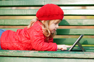 shutterstock / vitalinka - "happy smiling little girl working with a tablet pc and lying on a bench in a park" 